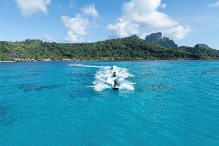 Two people jet skiing on blue ocean near a lush green island with hills.