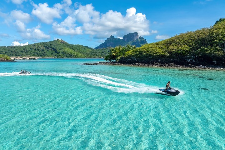 Two people jet skiing on turquoise water with lush hills and clouds in the background.