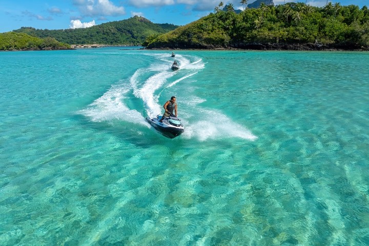 Two people riding jet skis on clear turquoise water with green islands in the background.