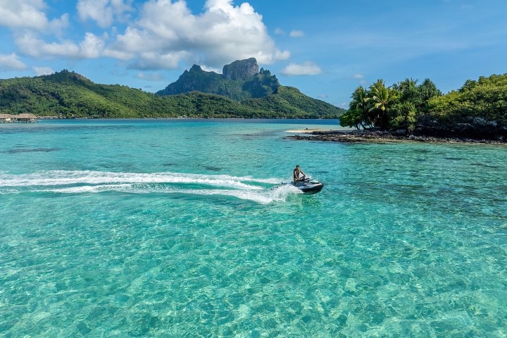 Person riding a jet ski on clear blue water near a green island with mountains.