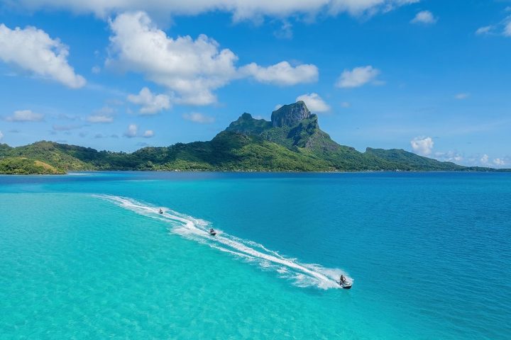 Blue sea with distant mountains and jet skis creating white trails under a clear sky.