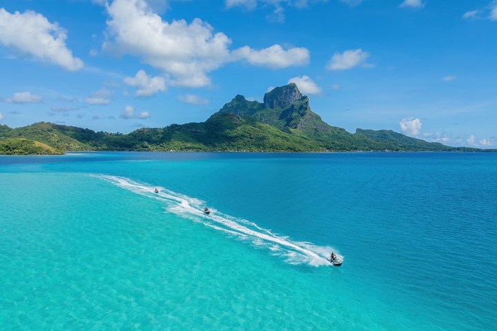 Blue sea with distant mountains and jet skis creating white trails under a clear sky.