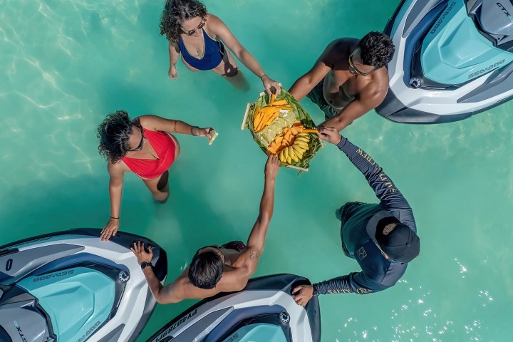 People in swimsuits on jet skis sharing tropical fruit in clear turquoise water.
