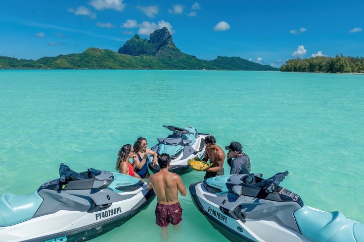 People on jet skis in clear turquoise water, with lush green island in the background.