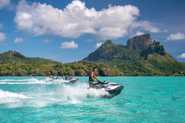 People riding jet skis on turquoise water with lush green mountains in the background.
