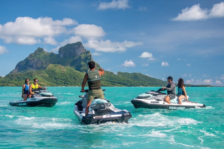 People on jet skis in turquoise water with lush green island and mountain in background.