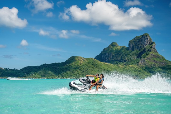 Two people on a jet ski in turquoise water with a mountain and blue sky in the background.