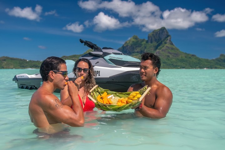 Three people in water near a jet ski, one holding a fruit platter, with a mountain in the background.
