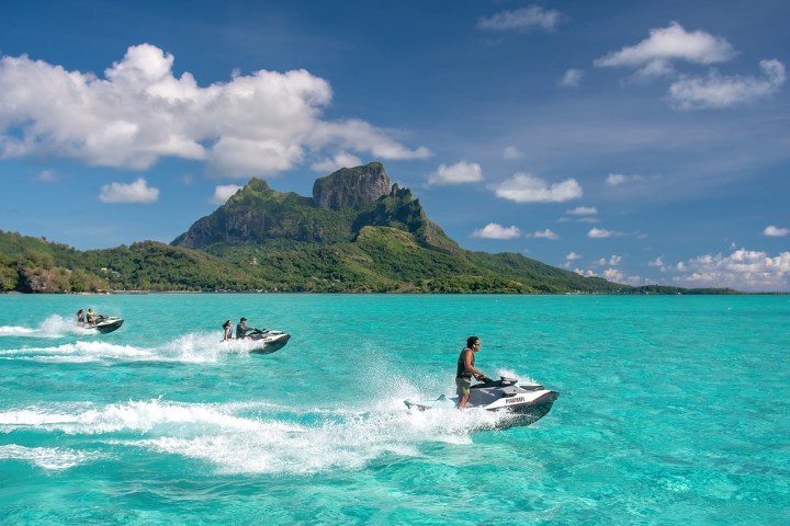 Three people on jet skis riding in turquoise water near a tropical island with lush mountains.