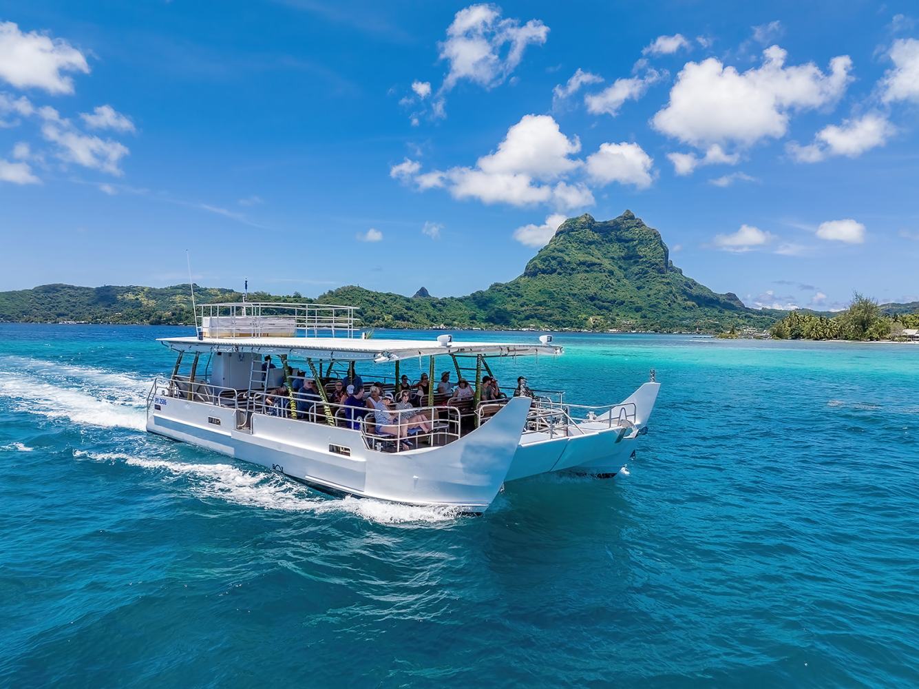 Tour boat on turquoise water near lush mountain under blue sky with clouds.