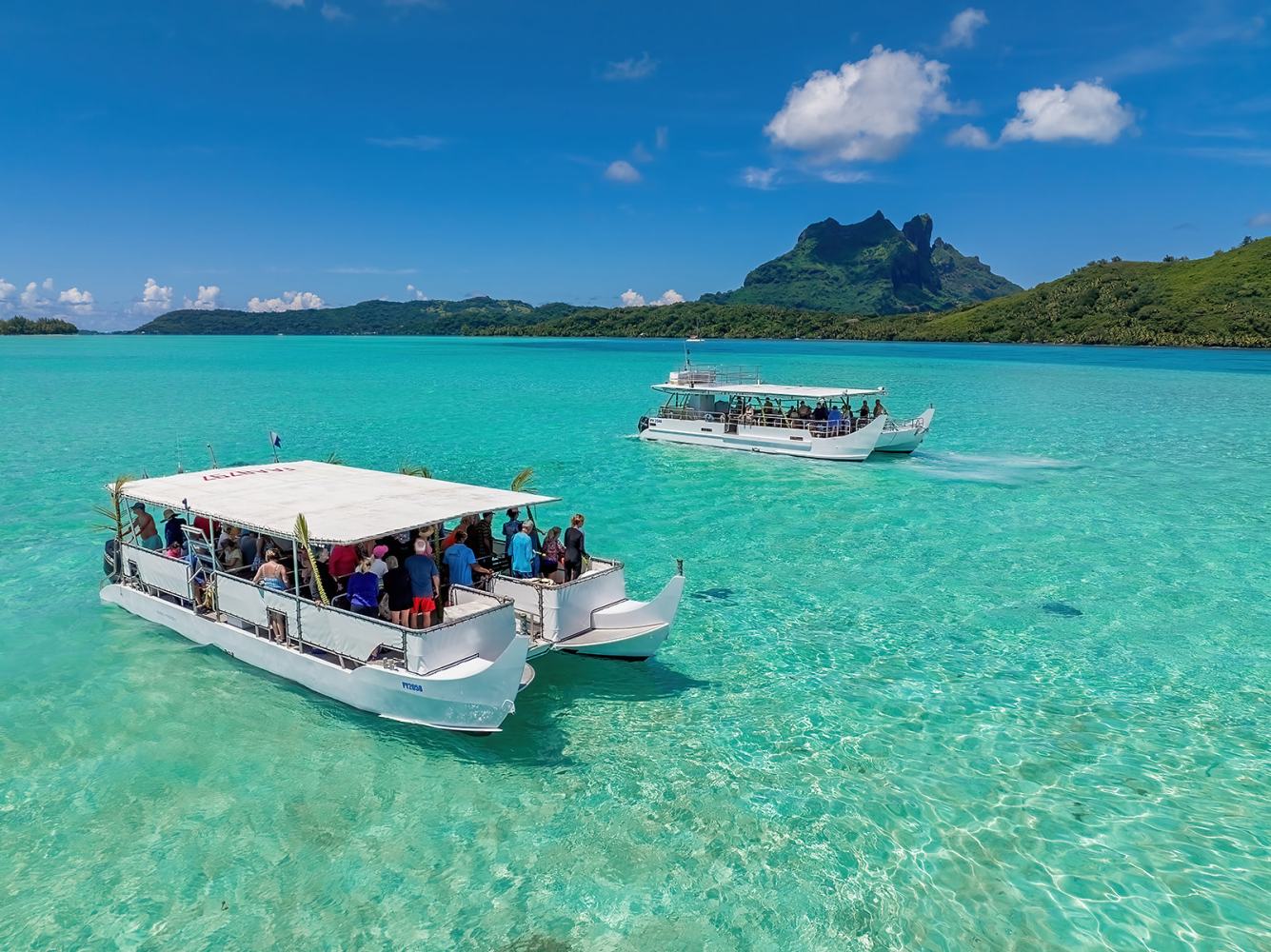 Two boats with tourists on clear turquoise water, mountains in background, and bright blue sky.