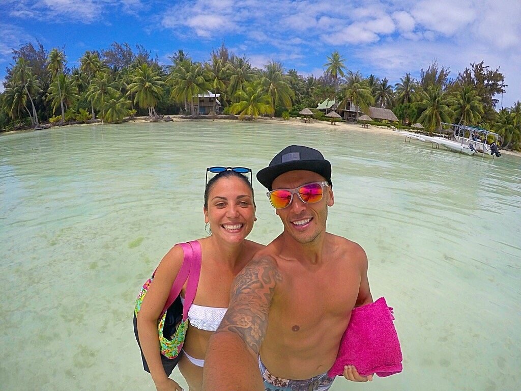 Smiling couple taking a selfie in shallow water with palm trees and huts in the background.